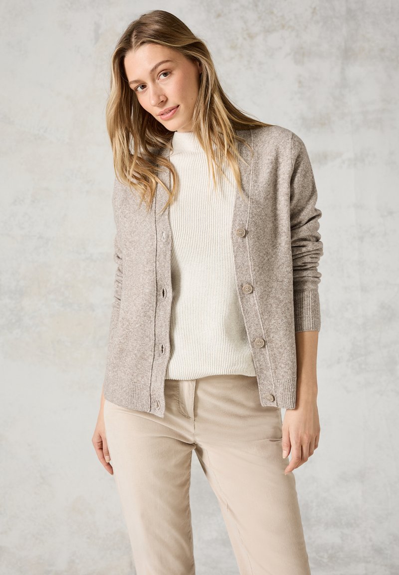 Cardigan beige chiaro realizzato in morbida lana, con una texture a coste, chiusura con bottoni e maniche lunghe, indossato sopra un maglione bianco e pantaloni beige.