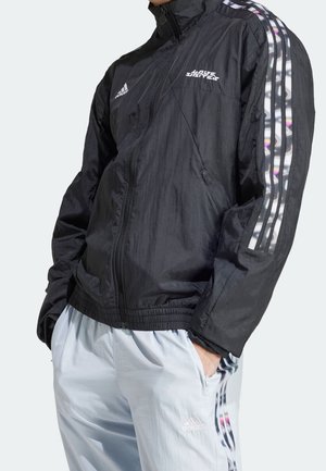 Person trägt eine schwarze Adidas-Jacke mit irisierenden Streifen an den Ärmeln und weiße Adidas-Hose mit passenden Streifendetails, Hände in den Taschen.