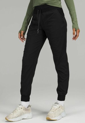 Persoon draagt zwarte joggingbroek, witte sokken, licht beige sneakers en een groen shirt met lange mouwen en duimgaten, staand tegen een effen achtergrond.