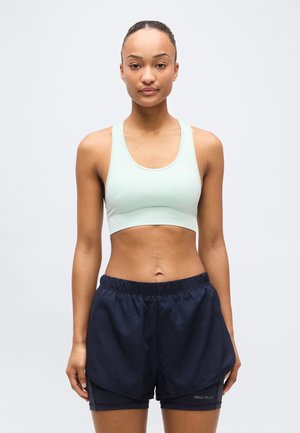 Femme debout, face à l'avant, portant un soutien-gorge de sport vert clair et un short de sport superposé bleu marine, sur un fond uni.