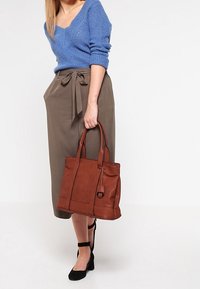 Bruine leren tote bag met dubbele handvatten, verticale naden en een gedeeltelijke klep-sluiting. Gecombineerd met een blauwe gebreide trui en een grijze rok.