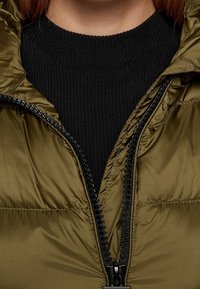 Olivově zelená péřová bunda se zapínáním na zip, pod níž je černý žebrovaný svetr. Hladká textura s výraznými švy.