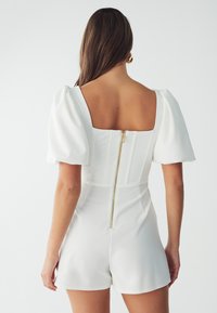 Witte rompert met pofmouwen, vierkante rug en verticale naden; beschikt over een verborgen ritssluiting met gouden hardware.