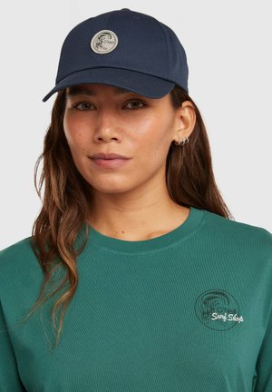 Tmavomodrá šiltovka s kruhovou nášivkou s logom O'Neill; má štruktúrovaný dizajn a zakrivený šilt. Nosená so zelenou košeľou s dlhými rukávmi.