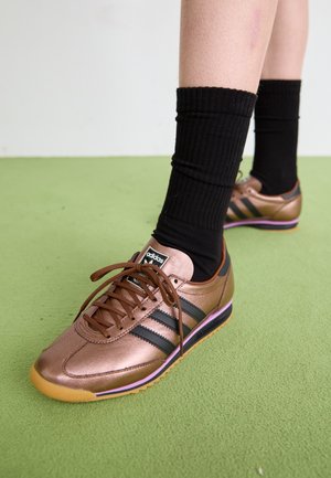 Μπρούντζινα αθλητικά παπούτσια με μαύρες ρίγες, καφέ κορδόνια και μοβ λεπτομέρεια στη σόλα. Συνδυάζονται με μαύρες κάλτσες με ραβδώσεις.