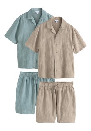 Jonge man draagt een taupekleurig, kort mouw knoophemd en bijpassende shorts, staand tegen een effen beige achtergrond met één hand in de zak.