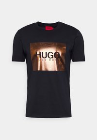 Schwarzes Baumwoll-T-Shirt mit einem rechteckigen bronzefarbenen Metallic-Druck, der den Schriftzug "HUGO HUGO BOSS" in fetten schwarzen Buchstaben zeigt. Kurzarm, Rundhalsausschnitt.