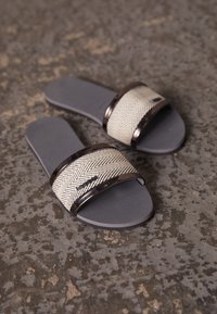 Un paio di sandali grigi a scivolo con suole testurizzate e cinghie in tessuto beige a spina di pesce accentuate da bordature scure, posizionati su un pavimento in pietra testurizzato.