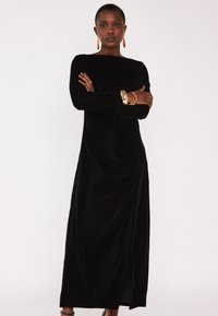 Robe en velours noire à manches longues avec une fente sur le côté, dotée d'un décolleté en scoop et de détails drapés. La texture brillante ajoute de la profondeur.