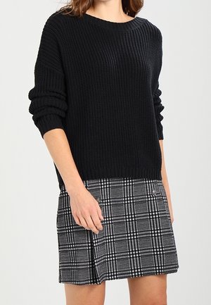 Vrouw die een zwarte geribbelde gebreide trui draagt met opgerolde mouwen en een zwart-witte geruite minirok.