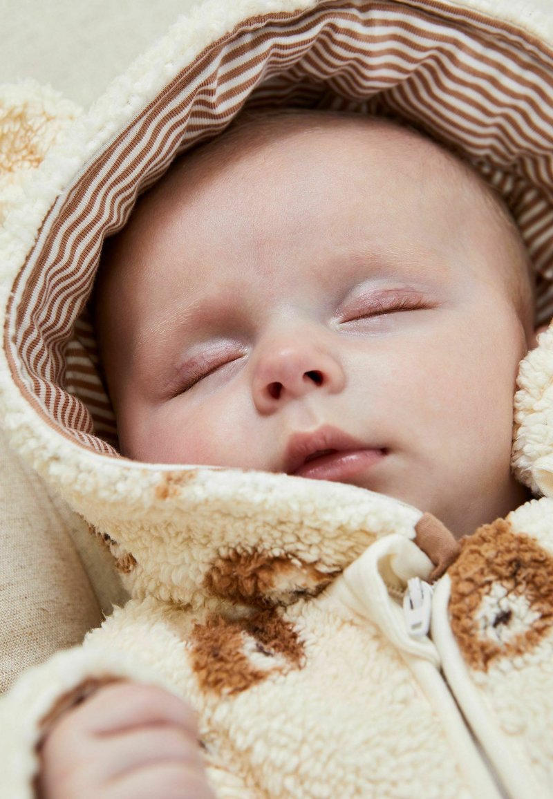 Bambino che dorme avvolto in un morbido abbigliamento con cappuccio in pile color crema e marrone, con foderatura a righe, mostrando occhi chiusi in modo pacifico e un viso rilassato.