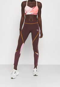 Il reggiseno sportivo presenta motivi geometrici rosa e arancioni; i leggings sono marrone scuro con accenti rosa e gialli. Entrambi sono aderenti e elasticizzati.