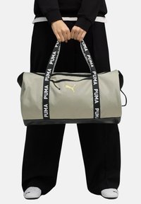 Bolsa de deporte gris con base negra, que presenta dos correas negras con el texto "PUMA" en blanco y un logo amarillo en la parte frontal. Cierre superior con cremallera.