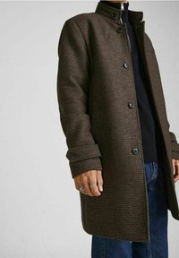 Homme portant un manteau boutonné à motif marron sur un sweat à fermeture éclair noir et un jean bleu, montrant des bagues à la main gauche.