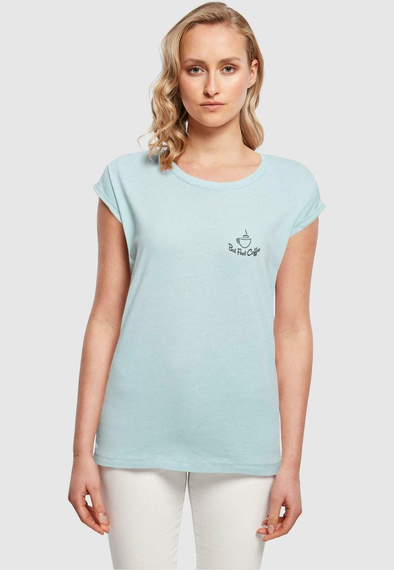 Lys blå t-shirt lavet af blødt stof, med rund halsudskæring og rullede korte ærmer. Den har et grafisk billede af en sort kaffekop og tekst.