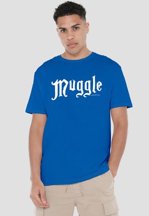 Young man wearing a blue T-shirt with "Muggle" printed in white gothic font, beige cargo shorts, standing with one hand in pocket.