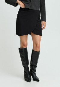 Jupe portefeuille noire avec une texture lisse, associée à des bottes en cuir noires mi-mollet avec des pointes et une finition élégante.