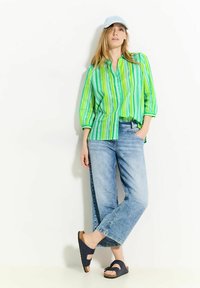 Camisa de botones a rayas verdes y azules, gorra azul clara, jeans anchos de mezclilla desgastados, y sandalias oscuras tipo slip-on con acentos de corcho.