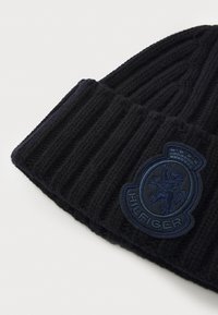 Beanie a coste nera con risvolto e una patch ricamata blu che presenta un simbolo di leone e il testo "HILFIGER".
