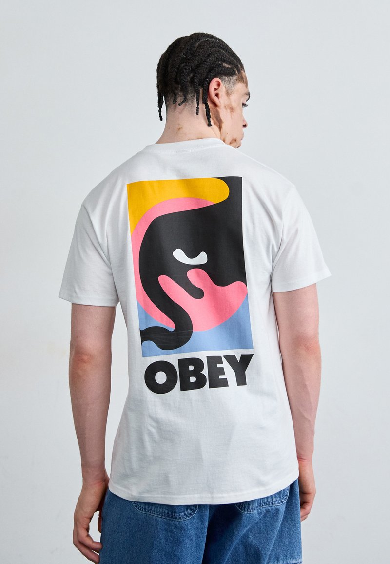 Fehér pamut póló, nagy grafikával a hátsó részen, merész fekete és színes absztrakt formákkal, valamint a "OBEY" szóval fekete színben.