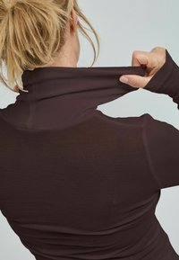 Mörk vinröd långärmad tröja med hög krage. Slätt, elastiskt tyg och figurnära design. Händer som drar upp kragen för att justera.