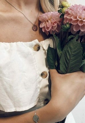 Weißes Leinenoberteil mit goldenen Knöpfen, silberne Kettenhalskette mit pinkem Anhänger und pinke Dahlienblüten, die mit grünen Blättern in der Hand gehalten werden.