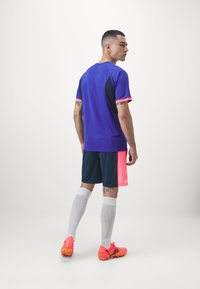 Lila Sportshirt mit pinken Akzenten, kombiniert mit marineblauen Shorts, die ein pinkes Seitenpanel haben. Weiße Socken und leuchtend pinke Schuhe runden das Outfit ab.