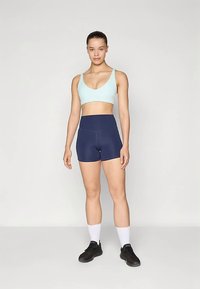Hellblauer Sport-BH kombiniert mit marineblauen, hoch taillierten Shorts. Modell trägt schwarze Sportschuhe und weiße Knöchelsocken. Schlichtes Design.