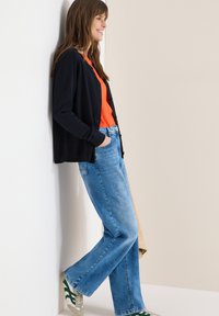 Femme appuyée contre un mur, souriante, portant un cardigan noir, un haut orange, un jean bleu et des baskets blanches avec des rayures vertes.