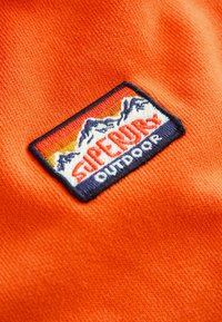 Žiarivo oranžová tkanina s textúrovaným povrchom, na ktorej je vyšitá nášivka. Nášivka má hory a text "SUPERDRY OUTDOOR."