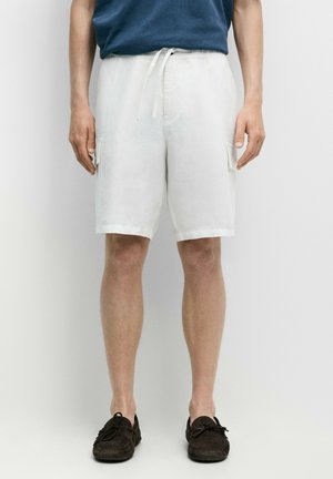 Mann trägt weiße Shorts mit Kordelzug und Seitentaschen, ein blaues Hemd, das hineingesteckt ist, und dunkelbraune Mokassins, steht vor weißem Hintergrund.