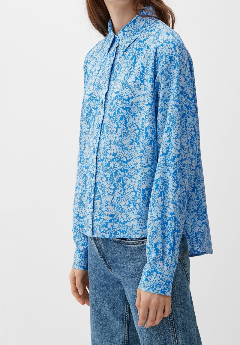 Femme portant une chemise bleue à fleurs à manches longues et un jean en denim bleu clair, debout devant un fond uni.