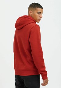 Red hoodie with a drawstring, textured fabric, ribbed cuffs, and hem. Worn with black pants, viewed from the back at an angle.