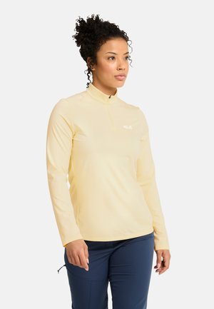 Donna con capelli ricci che indossa una maglia zip a maniche lunghe giallo pallido e pantaloni blu navy, in piedi davanti a uno sfondo bianco semplice.