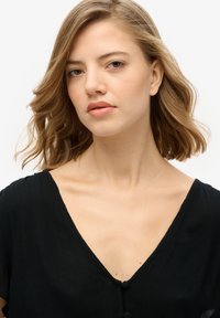 Blusa negra de manga corta con escote en V y detalles de botones. Tela suave y texturizada. Modelo con cabello ondulado de longitud media y maquillaje neutro.
