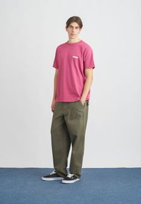 Ροζ βαμβακερό μπλουζάκι με μικρό λευκό λογότυπο, συνδυασμένο με χαλαρά ελαιοπράσινα παντελόνια και μαύρα αθλητικά παπούτσια σε μπλε επιφάνεια.