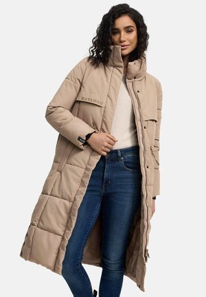 Lange beige pufferjas met een hoge kraag, voorzien van gestikte panelen, een ritssluiting aan de voorkant en branding op de zak; gedragen over een lichte trui en denim.