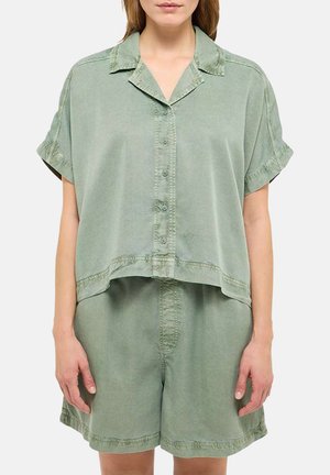 Femme portant une chemise verte sauge ample avec des boutons et des manches courtes, ainsi qu'un short décontracté assorti, debout les bras détendus.