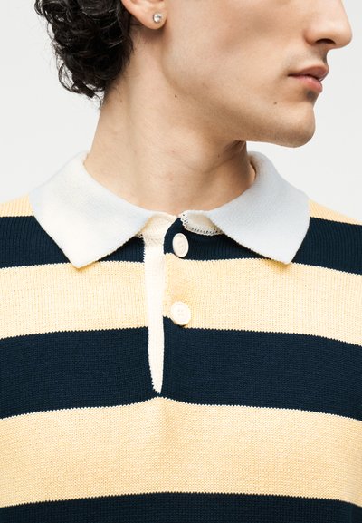 Personne portant un polo en maille à rayures horizontales bleu marine et beige avec un col blanc à deux boutons, la tête tournée de côté.