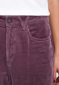 Pantalones de pana morados con prominentes rayas verticales, cierre de botón y detalle de bolsillo frontal. Textura suave y diseño informal.