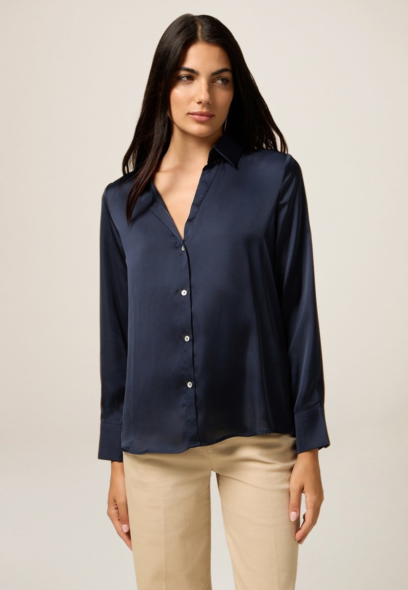Camisa de satén azul marino con botones, de manga larga, con cuello y dobladillo recto. Presenta botones similares a perlas en la parte delantera. Textura suave y lisa.
