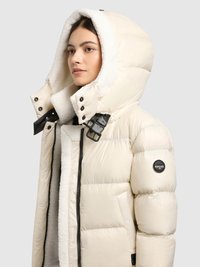 Kurtka puchowa w kremowym kolorze, z dużym kapturem wykończonym futerkiem, czarnymi elementami, pikowanym wzorem oraz naszywką z logo na rękawie.