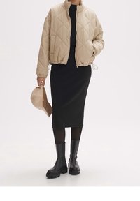 Osoba w beżowej pikowanej kurtce, czarnej midi sukience z prążkowanego materiału, czarnych rajstopach, masywnych czarnych botkach, trzymająca beżową czapkę bejsbolową.