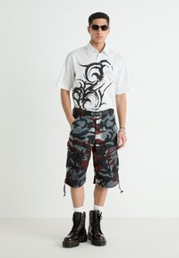 Camisa blanca de manga corta con diseño negro en espiral, combinada con pantalones cortos cargo de camuflaje negro, rojo y gris. Botas de combate negras completan el look.