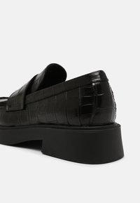 Mocassin en cuir noir avec texture crocodile et semelle plate-forme épaisse noire, vu en gros plan sous un angle arrière sur fond blanc.