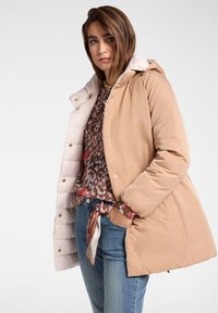 Béžový zimní kabát s lesklým, světlým vnitřkem, vybavený kapucí, knoflíky a bočními kapsami, nošený přes vzorovanou halenku a džíny.