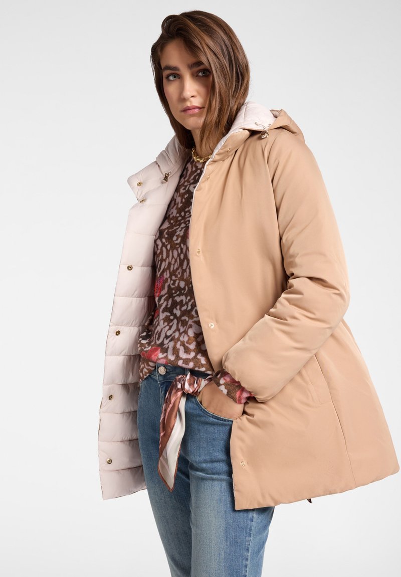 Béžový zimní kabát s lesklým, světlým vnitřkem, vybavený kapucí, knoflíky a bočními kapsami, nošený přes vzorovanou halenku a dž�íny.