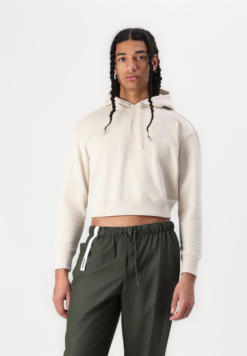 Cropped beige hoodie in soft fabric with a drawstring hood, featuring ribbed cuffs. Paired with dark green drawstring pants with side accents.