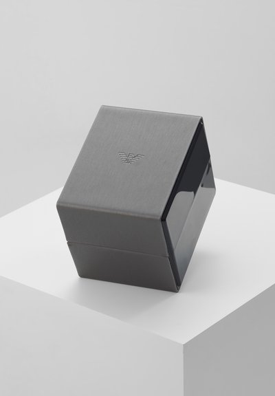 Boîte mate grise de forme cubique avec le logo Armani sur le dessus, placée sur un socle blanc sur un fond gris clair.