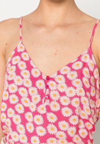 Top sin mangas rosa con un patrón de margaritas blancas, presenta un escote en V y tres botones rosas en la parte delantera, hecho de una tela ligera.